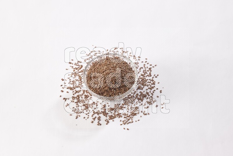 A glass bowl full of flax seeds surrounded by flax seeds on a white flooring