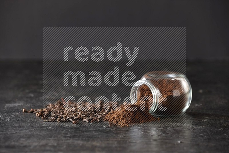 A flipped glass spice jar full of cloves powder and powder came out of it with cloves spread on textured black flooring