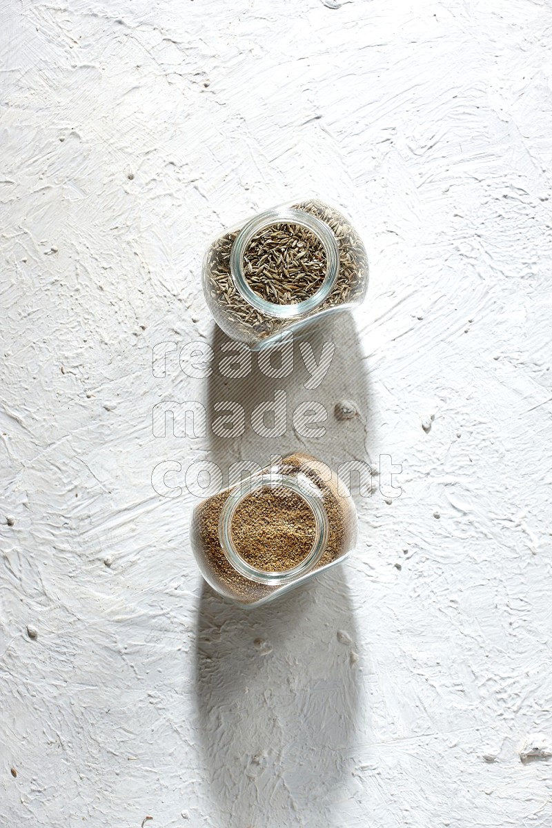 2 glass spice jars full of cumin powder and cumin seeds on textured white flooring