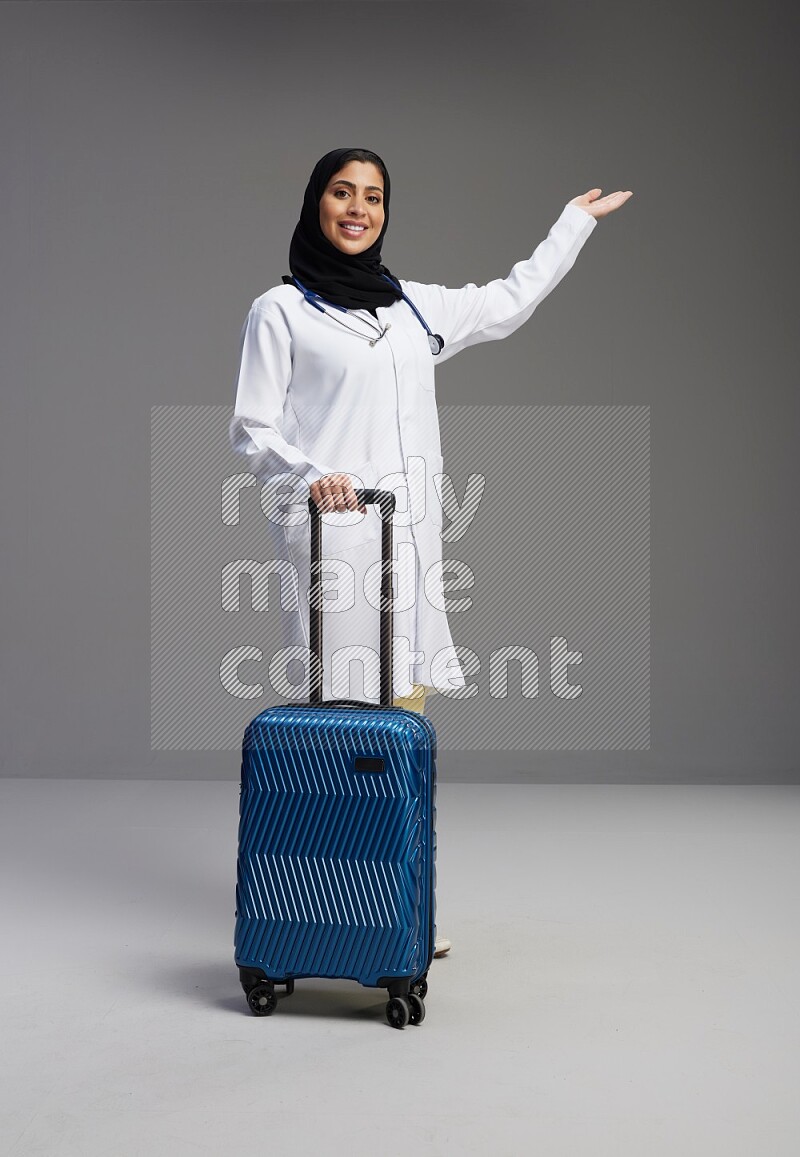Saudi woman wearing lab coat with stethoscope standing holding Travel bag on Gray background