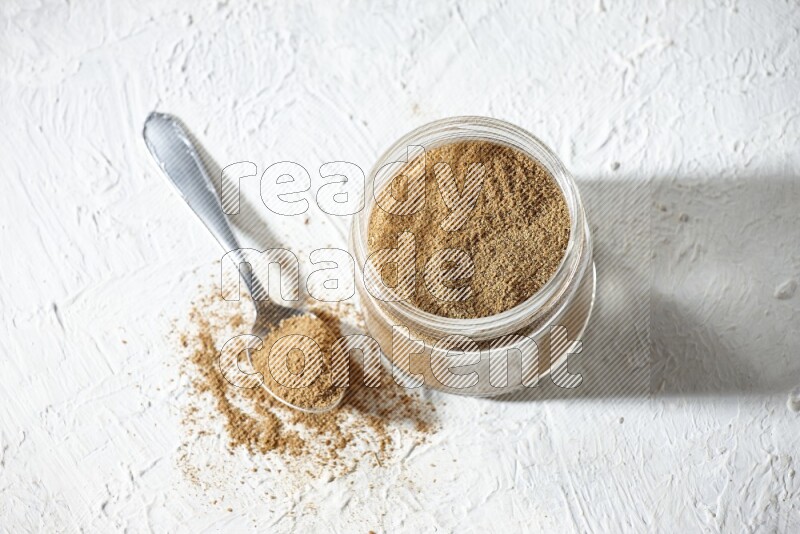 A glass jar and a metal spoon full of cumin powder on textured white flooring