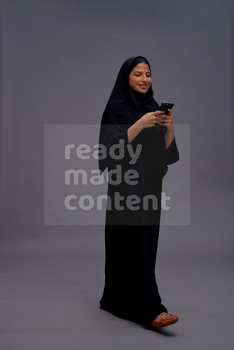 Saudi woman wearing Abaya standing texting on phone on gray background