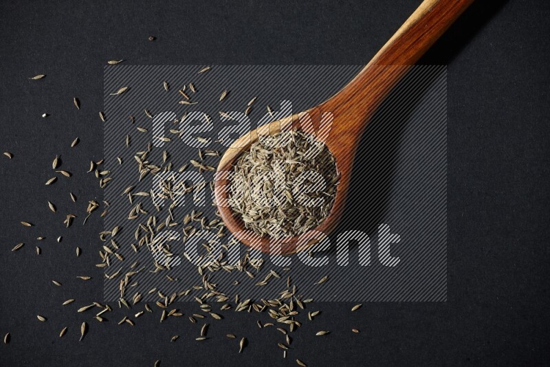 A wooden ladle full of cumin seeds on black flooring