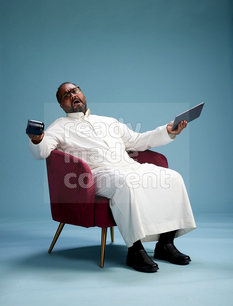 Saudi Man without shimag sitting on chair working on tablet on blue background