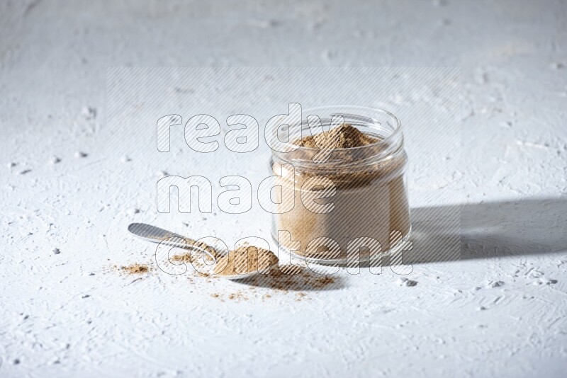 A glass jar and a metal spoon full of allspice powder on a textured white flooring
