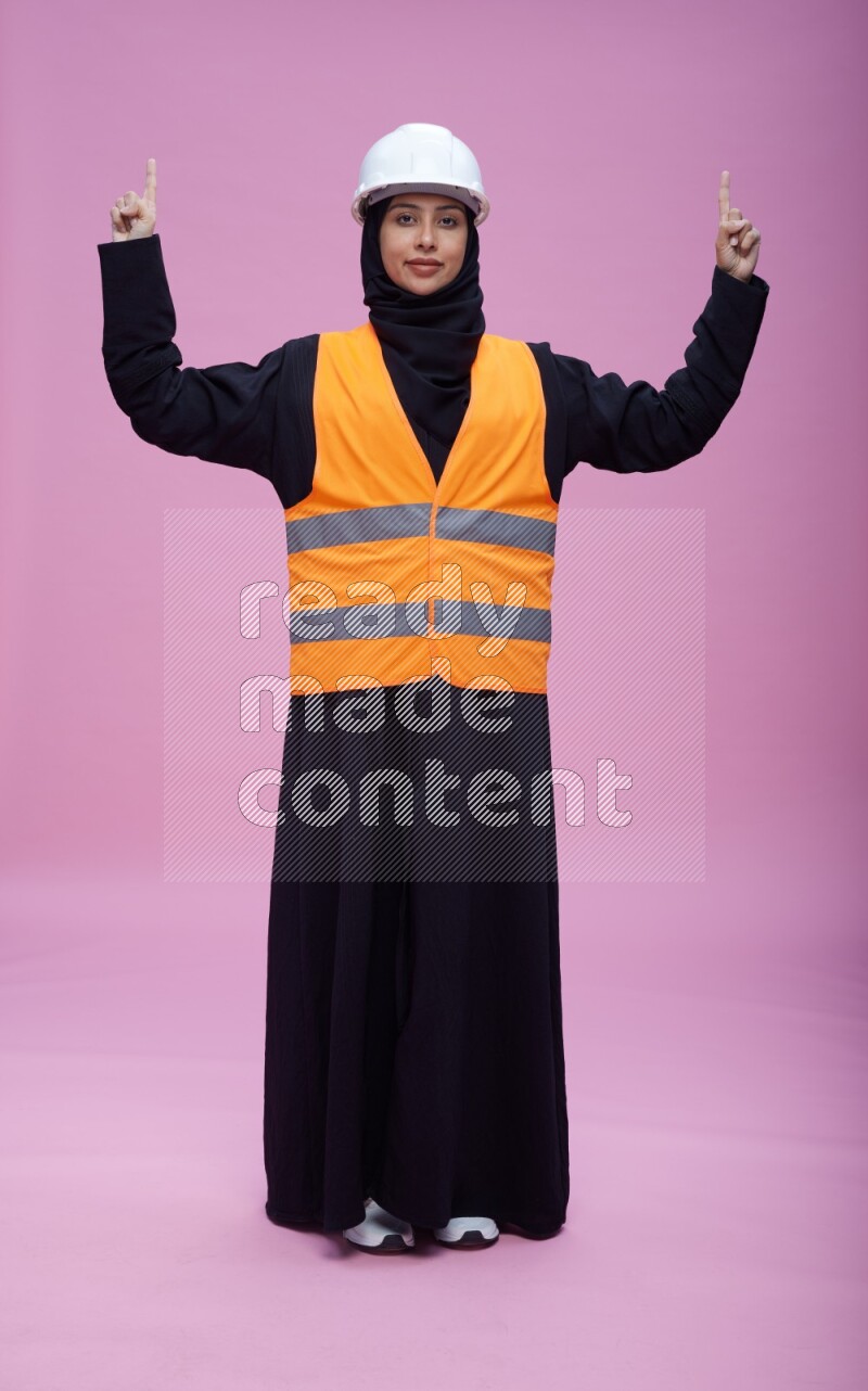 Saudi woman wearing Abaya with engineer vest and helmet standing interacting with the camera on pink background