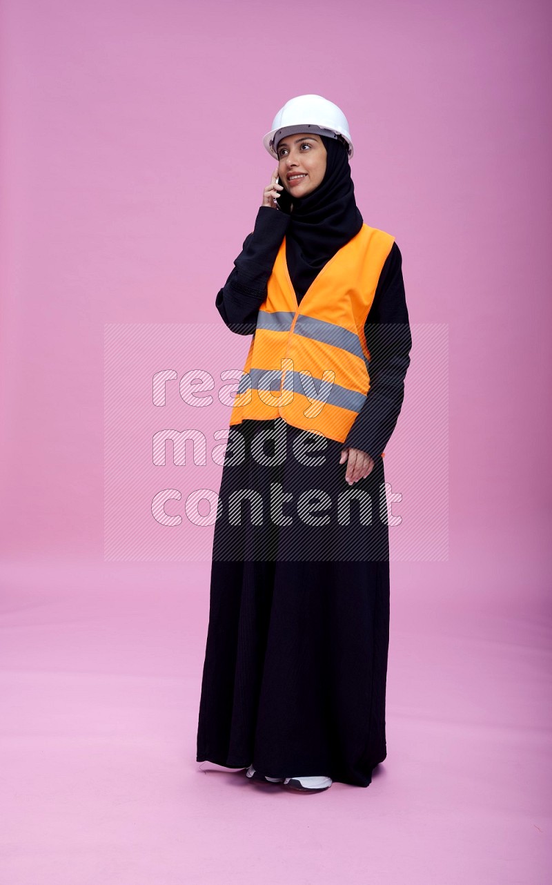 Saudi woman wearing Abaya with engineer vest and helmet standing talking on phone on pink background