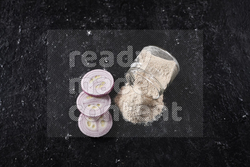 A glass jar full of onion powder flipped with some spilling powder on black background