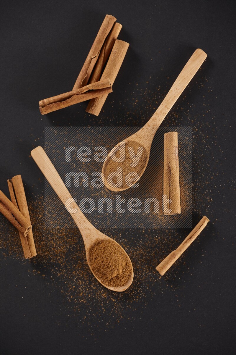 Cinnamon powder in two wooden spoons with cinnamon sticks on black background
