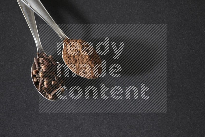2 metal spoons, one full of cloves grains and the other full of cloves powder on a black flooring