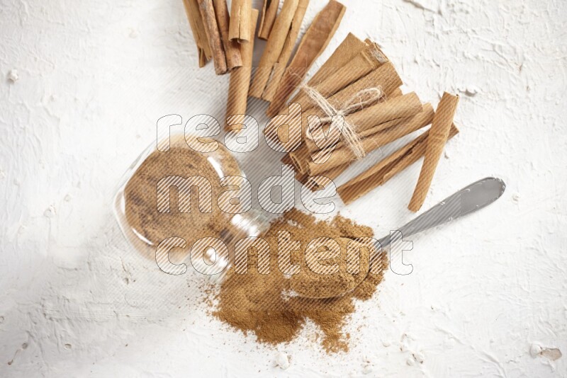 Flipped herbs glass jar full of cinnamon powder with a metal spoon full of powder and cinnamon sticks on a textured white background