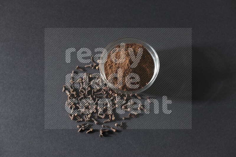 A glass bowl full of cloves powder with cloves grains spread on black flooring