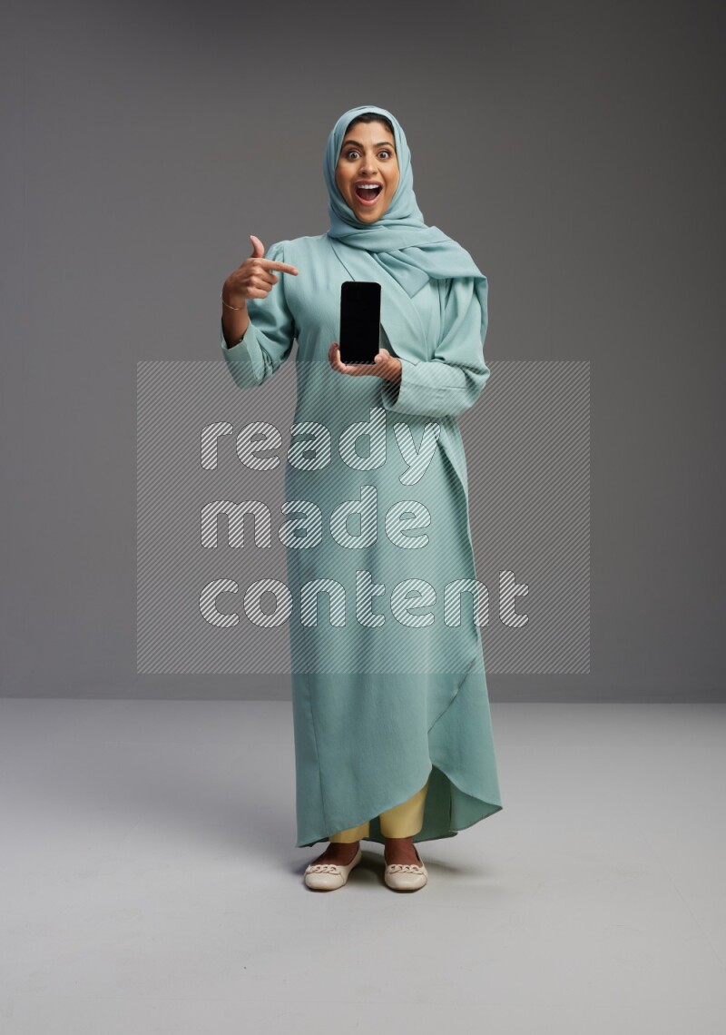 Saudi Woman wearing Abaya standing showing phone to camera on Gray background