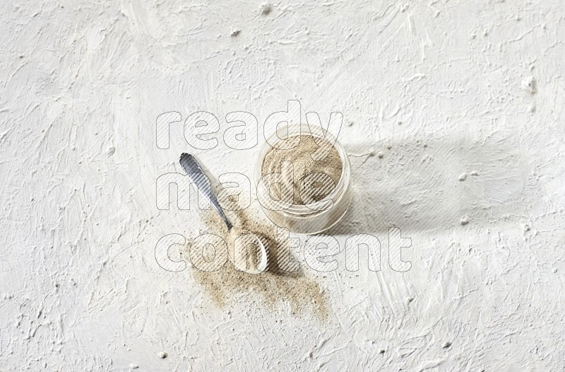 A glass jar and a metal spoon full of white pepper powder on textured white flooring