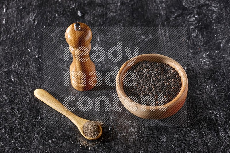 الفلفل الأسود في وعاء وملعقة خشبية مع مطحنة خشبية علي خلفية سوداء