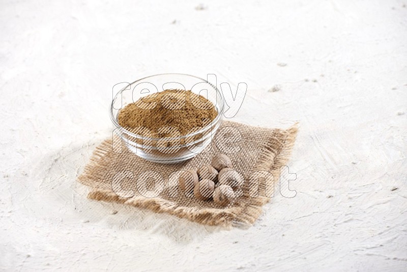 A glass bowl full of nutmeg powder with the seeds beside it on burlap fabric on a textured white flooring in different angles