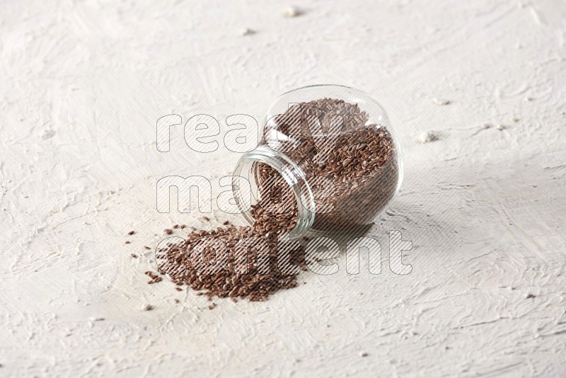 A glass spice jar full of flax seeds flipped and seeds spread out on a textured white flooring