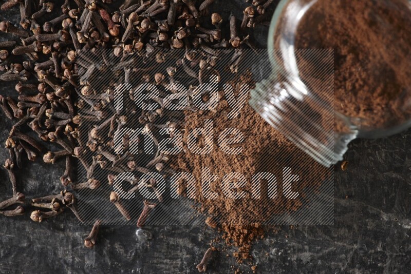 A flipped glass spice jar full of cloves powder and powder came out of it with cloves spread on textured black flooring