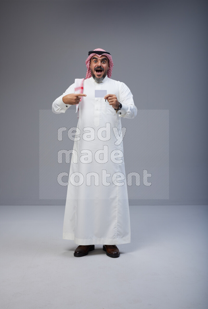 Saudi man Wearing Thob and red Shomag standing holding ATM card on Gray background