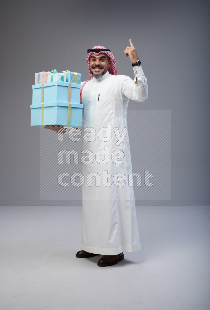 Saudi man Wearing Thob and red Shomag standing holding gift box on Gray background