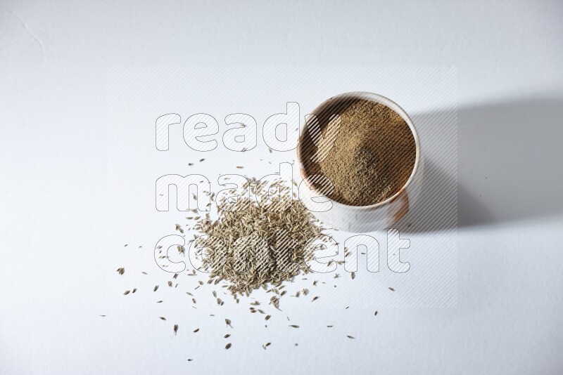 A beige bowl full of cumin powder with cumin seeds beneath it the bowl on a white flooring