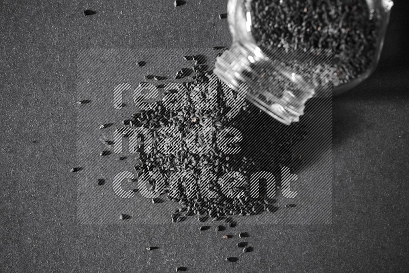 A flipped glass spice jar full of black seeds and the seeds spread out on a black flooring