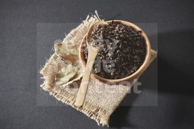 A wooden bowl, a wooden spoon full of cloves, and bay leaves (laurel) on a piece of burlap on a black flooring