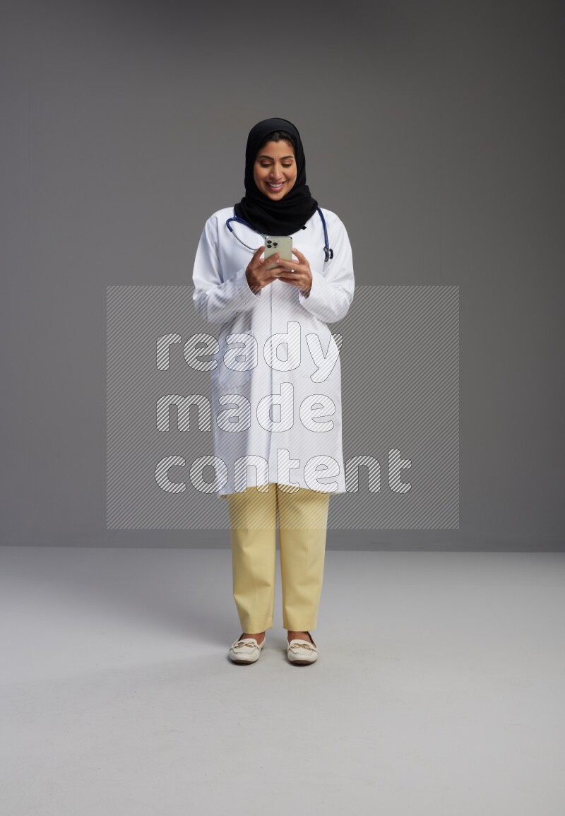 Saudi woman wearing lab coat with stethoscope standing texting on phone on Gray background