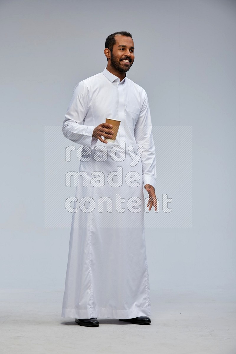 Saudi man wearing thob standing holding paper cup on gray background