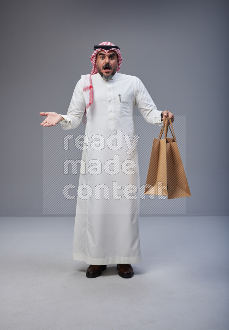 Saudi man Wearing Thob and red Shomag standing holding shopping bag on Gray background