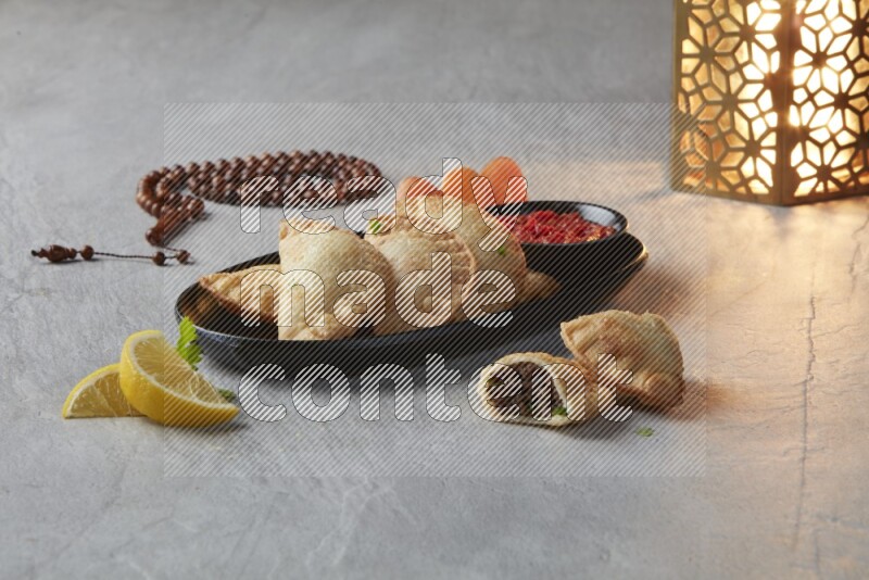 Five fried sambosas in an oval shaped black plate, beside a cut meat sambosa, a brown misbaha and a golden lantern on a gray background