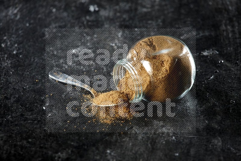 A flipped glass spice jar and a metal spoon full of cumin powder and powder spilled out on a textured black flooring