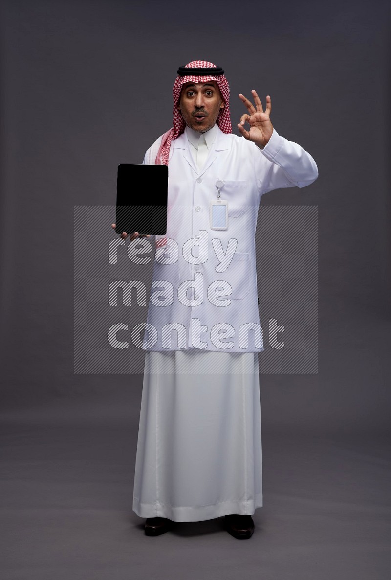 Saudi man wearing thob with lab coat and shomag with pocket employee badge standing showing tablet to camera on gray background