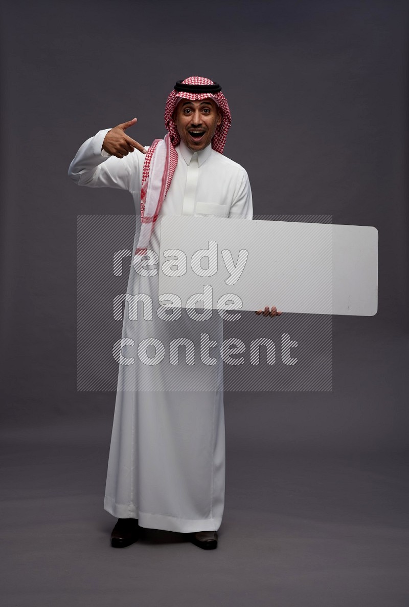 Saudi man wearing thob and shomag standing holding board on gray background