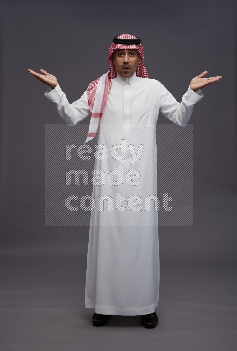 Saudi man wearing thob and shomag standing interacting with the camera on gray background