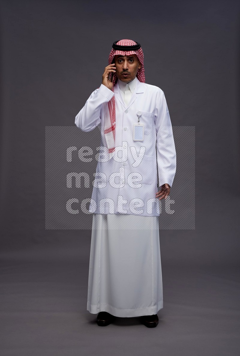 Saudi man wearing thob with lab coat and shomag with pocket employee badge standing talking on phone on gray background