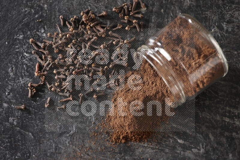 A flipped glass jar full of cloves powder with cloves spread on a textured black flooring