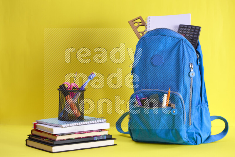 A school bag with assorted school supplies in and beside it on yellow background