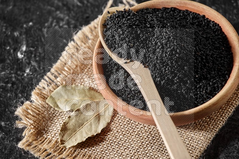 A wooden bowl full of black seeds with wooden spoon full of the seeds on it on burlap fabric on a textured black flooring in different angles