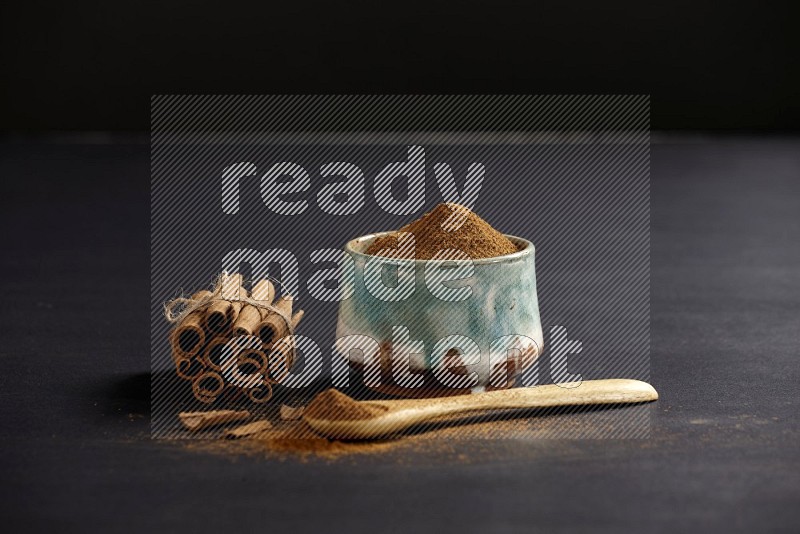 Ceramic bowl full of cinnamon powder and a wooden spoon full of powder with cinnamon sticks stacked and bounded on black background