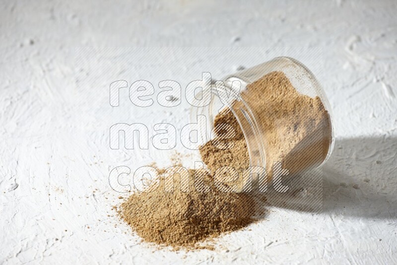 A flipped glass jar full of cumin powder and powder spilled out on textured white flooring