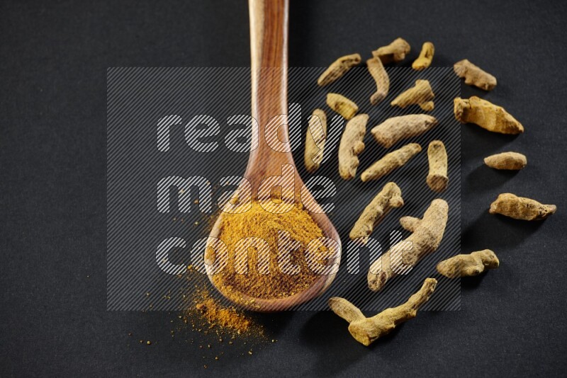 A wooden ladle full of turmeric powder with dried turmeric fingers on black flooring