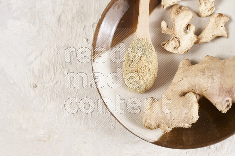 A wooden spoon full of ground ginger powder with fresh and dried ginger, all on a pottery plate on white background