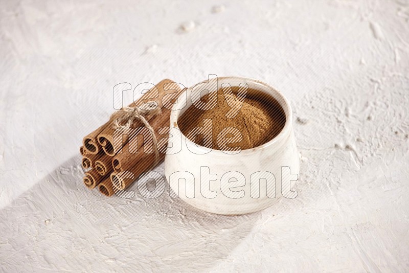 Cinnamon sticks stacked and bounded beside a beige bowl full of cinnamon powder on white background
