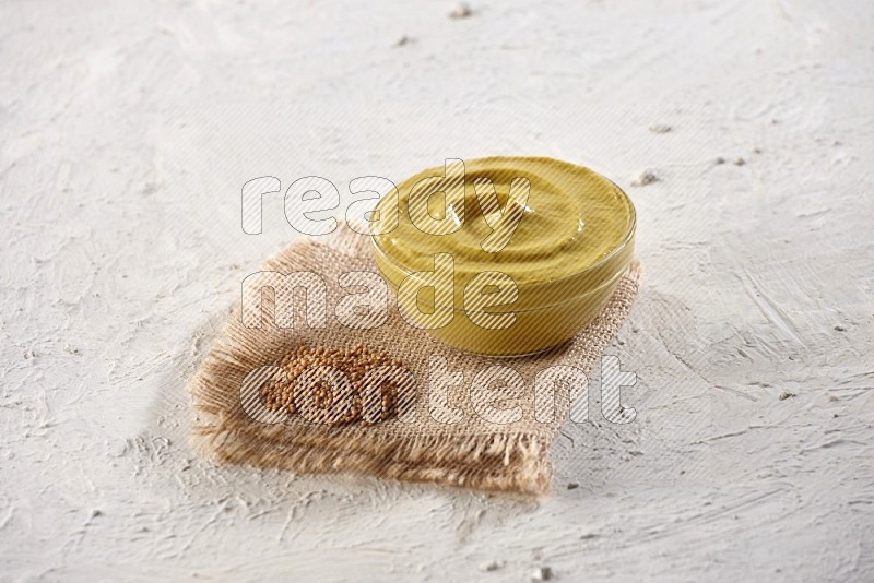 A glass bowl full of mustard paste set on a burlap piece with some mustard seeds spread on a textured white flooring