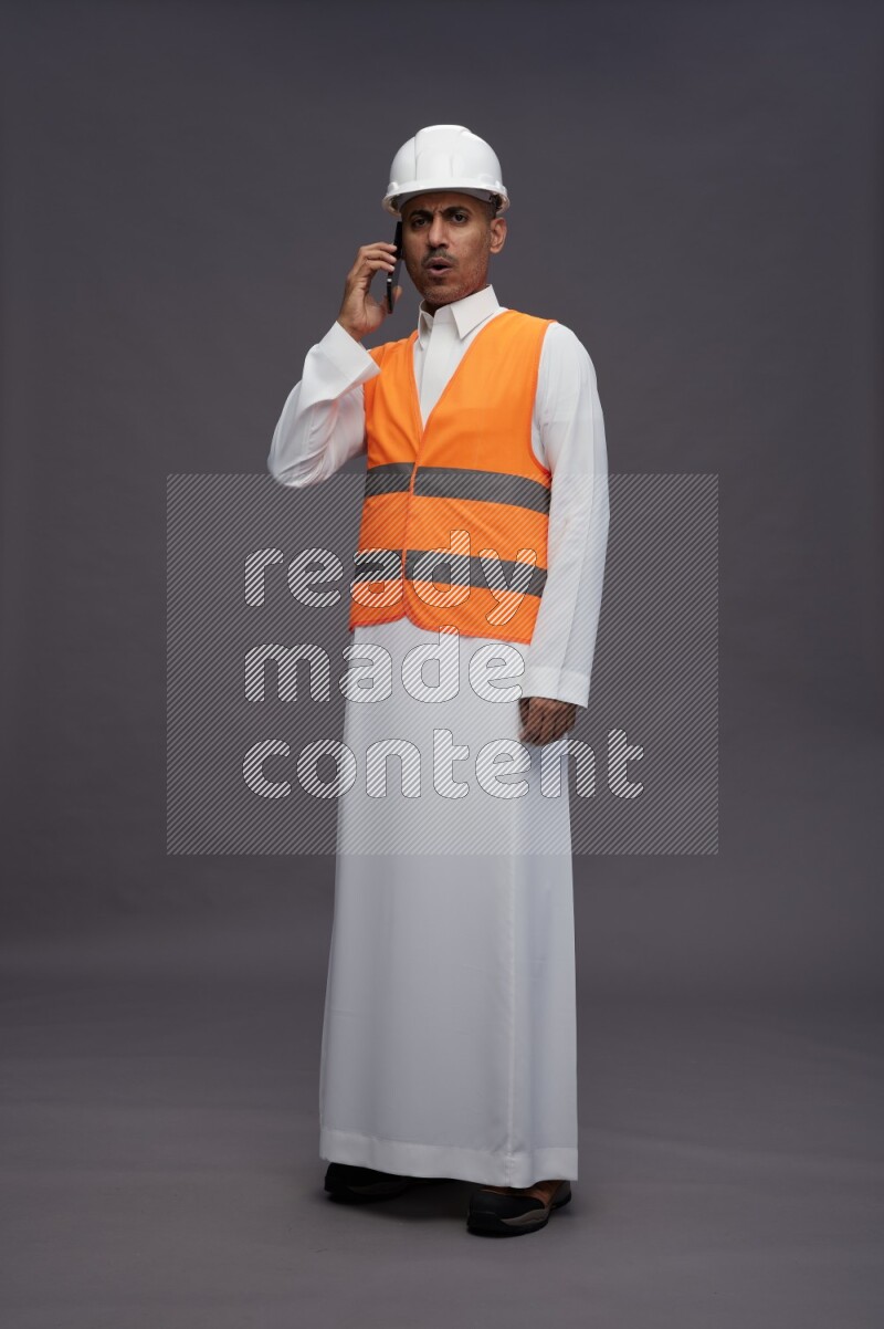 Saudi man wearing thob with engineer vest standing talking on phone on gray background