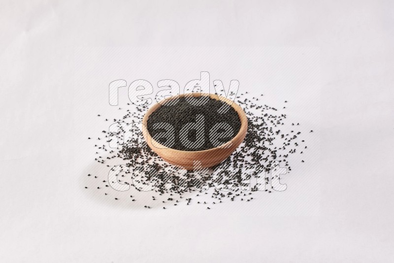 A wooden bowl full of black seeds and more seeds spread on a white flooring