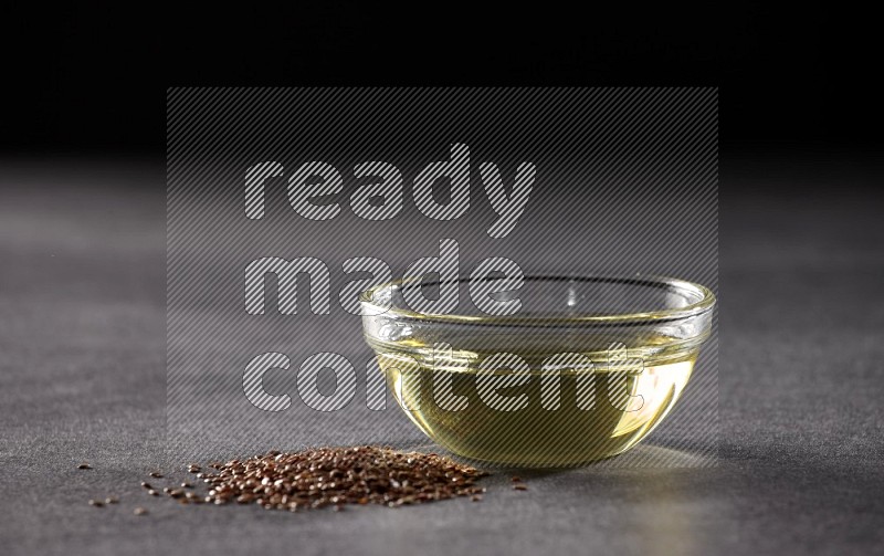 A glass bowl full of flaxseeds oil with some of flaxseeds beside it on a black flooring