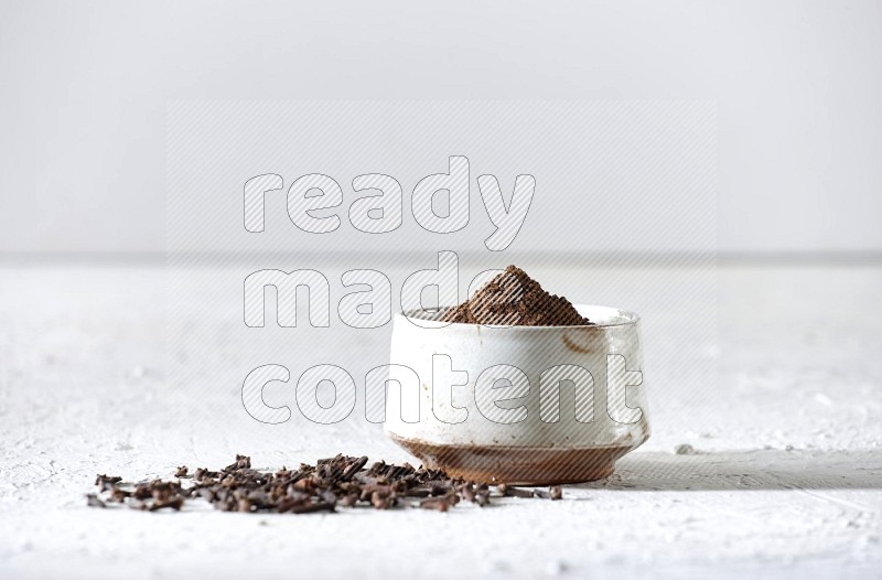 A beige ceramic bowl full of cloves powder and whole cloves on a white flooring