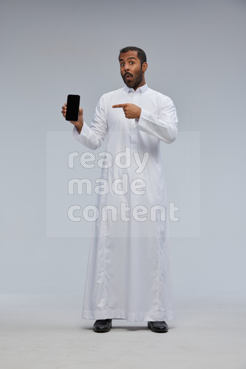 Saudi man wearing Thob standing showing phone to camera on Gray background